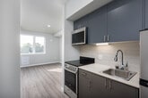 This single-floor unit showcases a clean, modern design with light wood-look flooring and neutral walls throughout, creating a cohesive and airy atmosphere. The galley-style kitchen is efficiently laid out with sleek gray cabinetry, light countertops, a tile backsplash, and stainless steel appliances, including an electric range, over-the-range microwave, and refrigerator. Under-cabinet lighting adds functionality to the workspace, which also features a single-basin sink with a contemporary faucet.

A dedicated laundry closet houses a stacked washer and dryer unit, providing convenient in-unit laundry without occupying significant space. The bathroom is designed with accessibility in mind, featuring grab bars, a gray vanity with storage, and light wood-look flooring that matches the rest of the unit.

The living area benefits from a large double-pane window that allows generous natural light to fill the room while offering outdoor views of the surrounding neighborhood. The overall layout is compact yet functional, with a straightforward flow connecting the kitchen corridor to the main living space. The unit's contemporary finishes and practical amenities make it a well-appointed and efficiently designed living space.