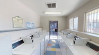 This shared laundry facility is a single-floor space designed with functionality and convenience in mind. The room is lined on both sides with rows of top-loading washing machines and dryers, providing ample capacity to serve multiple users simultaneously. The machines are arranged in a symmetrical layout, creating a clear central walkway that runs the full length of the room and leads to a glass-paneled security door at the far end, allowing for easy access and natural light to filter in.

The flooring features a distinctive colorful tile pattern with blue, yellow, orange, and white squares, adding a cheerful character to the utilitarian space. Overhead, fluorescent lighting ensures the room is well-lit throughout, supplemented by a window on the right-hand wall that brings in additional natural light and ventilation. A ceiling vent contributes to airflow and climate control within the space.

The walls are finished in a clean, neutral tone, maintaining a bright and sanitary atmosphere. Instructional and informational posters are mounted near the entrance, offering guidance on machine operation. Overall, the facility is thoughtfully organized, clean, and well-maintained, providing a practical and comfortable environment for laundry needs.