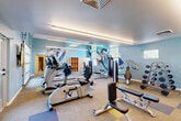 Located at 7845 Allison Way in Arvada, Colorado, this fitness center offers a comprehensive workout facility across a single floor. The space is divided into two distinct exercise rooms, each designed to support a variety of training needs.

The main exercise room is finished with a cool blue-toned color palette and features a vibrant athletic mural as a focal point, adding energy to the space. Linear LED lighting runs along the ceiling, providing bright, even illumination throughout. Carpet tile flooring in a mix of neutral, blue, and yellow tones defines workout zones across the room. This area is equipped with a full lineup of TRUE-brand cardio machines — including treadmills, ellipticals, and stationary bikes — as well as a cable machine, weight bench, and a full dumbbell rack. Large windows and a glass door offer views of an outdoor pool area, allowing ample natural light to filter in.

The second exercise room features warm hardwood flooring and light yellow walls, creating a distinct, inviting atmosphere. This studio-style space includes spin bikes, stability balls, TRX suspension trainers, and a wall-mounted flat-screen TV, making it ideal for group or functional fitness training. A window brings in natural light, keeping the room bright and open.

Together, the two rooms provide a well-rounded fitness environment suitable for cardio, strength, and functional training.