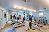 Located at 7845 Allison Way in Arvada, Colorado, this fitness center offers a comprehensive workout facility across a single floor. The space is divided into two distinct exercise rooms, each designed to support a variety of training needs.

The main exercise room is finished with a cool blue-toned color palette and features a vibrant athletic mural as a focal point, adding energy to the space. Linear LED lighting runs along the ceiling, providing bright, even illumination throughout. Carpet tile flooring in a mix of neutral, blue, and yellow tones defines workout zones across the room. This area is equipped with a full lineup of TRUE-brand cardio machines — including treadmills, ellipticals, and stationary bikes — as well as a cable machine, weight bench, and a full dumbbell rack. Large windows and a glass door offer views of an outdoor pool area, allowing ample natural light to filter in.

The second exercise room features warm hardwood flooring and light yellow walls, creating a distinct, inviting atmosphere. This studio-style space includes spin bikes, stability balls, TRX suspension trainers, and a wall-mounted flat-screen TV, making it ideal for group or functional fitness training. A window brings in natural light, keeping the room bright and open.

Together, the two rooms provide a well-rounded fitness environment suitable for cardio, strength, and functional training.