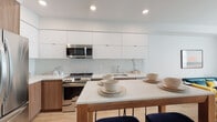 Located on the iconic Hollywood Boulevard in Los Angeles, this modern single-floor residence offers a thoughtfully designed layout with clean, contemporary finishes throughout. The open kitchen is a standout feature, showcasing flat-front white cabinetry extending to the ceiling, quartz countertops, a marble-look tile backsplash, and stainless steel appliances including a range and built-in microwave. A wood-framed dining table with a quartz surface serves as a natural gathering point between the kitchen and living areas. The living room is bright and welcoming, anchored by light hardwood flooring, a large window with a glass door that invites natural light, and a neutral palette that complements the space's modern aesthetic. A versatile secondary room with hardwood floors offers flexibility as a home office or lounge, complete with its own seating area. The bedroom is serene and airy, featuring a large frosted window that diffuses soft natural light across the light hardwood floors. The bathroom is sleek and minimal, with a backlit LED vanity mirror, quartz countertop, and undermount sink. Throughout the residence, crisp white walls and recessed lighting create a cohesive, polished ambiance.