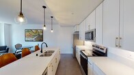 This well-appointed single-floor residence offers a thoughtfully designed layout that balances modern style with everyday functionality. The kitchen is a standout feature, showcasing crisp white cabinetry with brushed gold hardware, white quartz countertops, and a full suite of stainless steel appliances including a refrigerator, electric range, and microwave. A central island with seating for two is illuminated by a trio of globe pendant lights, creating an inviting space for casual dining or entertaining. The open-plan living area flows naturally from the kitchen and is bathed in natural light from large windows that frame impressive urban skyline views. The living space is anchored by sleek leather seating and contemporary furnishings, lending a sophisticated, minimalist feel. The bedroom is a serene retreat, featuring soft carpeting, recessed lighting, coordinated dark wood nightstands, and curated wall art. A closet provides convenient storage. The bathroom is finished with classic black-and-white hexagonal mosaic tile flooring and a full walk-in shower enclosed by sliding glass doors with white subway tile surround, complemented by a clean white vanity. Throughout the space, a neutral color palette and clean lines create a cohesive and refined urban living environment.