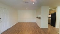 This single-floor space presents a straightforward and functional layout highlighted by warm-toned hardwood-style flooring that extends throughout the main living area, lending a sense of continuity and warmth to the interior. The walls are finished in a clean, neutral off-white tone, creating a bright and versatile backdrop that accommodates a wide range of furnishing styles and uses.

The ceiling features a textured finish and is equipped with a ceiling-mounted track lighting fixture, providing adjustable illumination across the space, complemented by a recessed downlight that adds focused ambient lighting. A central HVAC vent is also integrated into the ceiling, ensuring climate control throughout.

A notable architectural detail is the pass-through window cut into one wall, fitted with a small ledge or shelf beneath it. This feature suggests a connection to an adjacent room or kitchen area, adding a degree of openness and practicality to the layout. White baseboards frame the perimeter of the room cleanly, completing the overall polished, move-in-ready aesthetic. The space feels open, uncluttered, and well-suited for a variety of functional configurations.