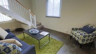 This two-storey space features a ground-floor living area characterized by polished concrete flooring that lends an industrial yet functional aesthetic to the room. A bright window fitted with white blinds allows natural light to fill the space, creating an open and airy atmosphere. The living area is anchored by a green shag rug layered over the concrete floor, complementing the neutral, cream-toned walls. A sleek, dark coffee table sits centrally, paired with a patterned accent chair and a blue sofa, offering a comfortable and practical seating arrangement. A carpeted staircase with a white-painted balustrade and warm timber handrail connects the ground floor to the upper level, adding architectural interest to the layout. The upper floor is visible from below via an open balustrade railing, contributing to a sense of vertical space and connectivity between the two levels. Overall, the space combines raw industrial elements with everyday comfort, making it a versatile and liveable environment suited to a range of occupants.