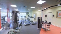 This fitness center offers a comprehensive workout environment designed to accommodate a range of exercise needs. The space is laid out on a single floor and benefits from a bright, welcoming atmosphere created by large recessed panel lighting in a drop ceiling overhead. Natural light filters in through windows along one wall, adding to the open and energizing feel of the room.

The cardio section is well-appointed, featuring multiple treadmills and an upright stationary bike, providing ample options for cardiovascular training. A wall-mounted television is positioned for convenient viewing during workouts. Complementing the cardio equipment, the strength training area includes a cable weight machine and a dedicated free weight bench, catering to users focused on resistance and muscle conditioning. An additional functional training station with a cable pull system rounds out the strength offerings.

The flooring consists of durable, dark rubberized tiles throughout most of the space, with a section of contrasting orange rubber matting designated for specific training activities. The neutral-toned walls and clean, organized layout contribute to a practical and professional atmosphere. A private room accessible via a glass-paneled door can be seen at the rear of the space, offering additional utility. Overall, this fitness center is thoughtfully arranged to maximize usability and comfort.