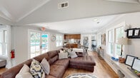 Located at 2201 Mossy Oaks Rd in Beaufort, SC, Bay South is a thoughtfully designed single-floor space that blends comfort and functionality. The entrance greets visitors with dramatic vaulted, beamed ceilings clad in white shiplap, floor-to-ceiling windows, and elegant French doors that flood the interior with natural light while offering views of the surrounding mature trees and landscaping.

The main living and lounge area features warm, rustic hardwood flooring throughout, complemented by soft neutral gray walls and recessed lighting. Comfortable seating arrangements and tasteful décor create an inviting atmosphere, while the open layout flows seamlessly into a dedicated workspace area, complete with a desk setup and wall-mounted display screen.

A separate kitchenette area is well-appointed with rich wood cabinetry, granite countertops, stainless steel appliances, and a sink — ideal for hosting or everyday convenience. High-top tables and bar stools provide casual seating adjacent to windows overlooking an outdoor pool area. Crown molding and clean architectural details add a polished finish throughout.

The space also includes a coffee and refreshment station with direct access to the exterior, enhancing its versatility. Overall, Bay South offers a bright, professional, and welcoming environment suited for a variety of uses.
