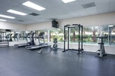 Located at 3200-3205 Northwood Dr in Concord, California, this well-equipped fitness center offers a generous single-floor layout designed to support a comprehensive workout experience. The main exercise area is finished with durable rubber flooring and illuminated by recessed panel lighting set within a drop-tile ceiling, creating a clean, bright environment conducive to focused training.

The strength training section is stocked with a variety of Life Fitness cable machines, multi-use weight stations, adjustable benches, a power rack, and a dumbbell rack, providing ample options for both resistance and functional training. Full-length mirrors line the walls, enhancing the sense of space while allowing users to monitor their form throughout their workout.

On the cardio side, multiple treadmills and a stair climber are positioned along a bank of windows that look out onto lush, tree-lined outdoor scenery, offering natural light and pleasant views during exercise. A wall-mounted television is also present for added convenience.

Toward the rear of the facility, a dedicated support area houses rows of gray metal lockers for personal storage, along with clearly marked restroom facilities and a water dispenser. The overall layout is open and thoughtfully organized, ensuring ease of movement between equipment zones. The space presents a functional and accessible fitness environment suitable for a wide range of users.