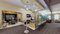 This well-appointed single-floor community space is designed to accommodate a variety of social and functional needs. The centerpiece of the main area is a large bar with a dark granite countertop, chrome accents, and pendant lighting overhead, paired with bar-height seating along one side. A wall-mounted television and built-in cabinetry complete the bar setup, while an adjacent dining area provides additional seating for gatherings.

Multiple lounge zones feature plush sofas, upholstered armchairs, and accent tables, all unified by a rich patterned carpet and a warm color palette of teal and cream. Vaulted and tray ceilings with cove lighting lend an elevated, airy feel throughout. A distinctive curved sunroom, lined with floor-to-ceiling arched windows and cream drapery, offers high-top bistro tables with views of an outdoor pool area, flooding the space with natural light.

Direct access to the pool is provided through glass-paned doors. The kitchen is fully equipped with dark espresso cabinetry, granite countertops, stainless steel appliances including a double wall oven, gas cooktop, built-in microwave, refrigerator, and dishwasher, and white tile flooring — ideal for catering or community events.