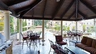 This outdoor pavilion offers a sophisticated resort-style retreat centered around a stunning pool area. The structure is defined by its soaring vaulted ceiling with rich, dark exposed wood beams, supported by classic white columns that frame open views of the surrounding grounds. The space is furnished with wrought-iron bar-height tables and chairs accented with deep red cushions, creating a refined yet relaxed atmosphere ideal for lounging and socializing. The polished concrete flooring adds a sleek, durable finish underfoot. Beyond the pavilion, the pool deck extends outward with additional seating areas shaded by umbrellas and lined with lounge chairs, inviting extended outdoor enjoyment. Lush tropical landscaping, including palm trees and tall cypress, frames the environment beautifully. In the background, Mediterranean-inspired multi-story buildings with warm stucco facades and terracotta rooflines create a cohesive architectural aesthetic. The open-air design allows natural light and breezes to flow freely throughout, blurring the line between sheltered comfort and the outdoors. This space is well-suited for gatherings, leisure, and al fresco dining in an upscale, resort-like setting.