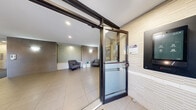 This single-floor lobby presents a well-designed, contemporary entrance space that combines functionality with modern aesthetics. The entry point features a secure glass sliding door with a keypad intercom system, providing controlled access to the interior. Large-format light beige tiles extend throughout the space, creating a clean, cohesive foundation underfoot.

The main lobby area is generously proportioned, with recessed pot lighting illuminating the space from above, complemented by wall-mounted sconce fixtures. Taupe and grey wall tile cladding lends a sophisticated, layered texture to key accent walls, while a central decorative panel with an organic, flowing relief pattern serves as a focal point. Two grey upholstered armchairs with a small side table create a comfortable waiting area, accented by subtle teal decorative accessories.

A dedicated mailroom area houses a full bank of secured resident mailboxes set against a textured tile wall, with a large window offering views of the surrounding greenery outside. An adjacent corridor leads to a secondary interior space featuring a bold teal accent wall, dark wood-panel storage units, and a slim white console table. A vibrant botanical artwork adds a pop of color to the overall neutral palette, enhancing the welcoming ambiance of the space.