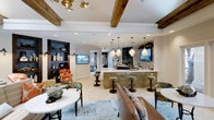 The Aqua Lounge is a thoughtfully designed single-floor gathering space that seamlessly blends rustic charm with contemporary elegance. Exposed wooden ceiling beams add warmth and architectural character, complemented by recessed lighting and a brass ceiling fan that enhance the inviting atmosphere. Decorative pendant lights with geometric, jewel-like shades hang above a central bar area, which features a gleaming white countertop, gold mosaic tile base, and a natural stone backsplash. Bar stools with colorful upholstery line the bar, while mounted televisions provide entertainment for guests.

The lounge area is furnished with a rich leather sofa, vibrant patterned accent chairs in warm oranges and teals, and a round marble-top cocktail table, all anchored by a blue vintage-style area rug. A built-in dark shelving unit along one wall displays decorative vases and accessories, adding a curated, boutique feel. The neutral tile flooring ties the space together, while the open layout encourages social interaction. The overall ambiance is sophisticated yet relaxed, making the Aqua Lounge an ideal setting for casual gatherings and social events.