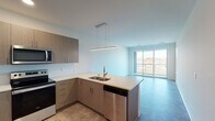 This single-floor unit showcases a clean, contemporary design with an open-concept layout that seamlessly connects the kitchen and living area. The kitchen is well-appointed with flat-front wood-tone cabinetry, white quartz countertops, a subway tile backsplash, and a full suite of stainless steel appliances, including an electric range, over-the-range microwave, dishwasher, and refrigerator. A peninsula with an undermount sink provides additional workspace and a natural division between the kitchen and the living area. A sleek linear pendant light fixture adds a modern touch overhead. The living area features durable tile-style flooring and is bathed in natural light from large sliding glass doors that open to a private balcony with open outdoor views of the surrounding neighborhood. The bedroom is carpeted for comfort and includes a generously sized walk-in closet with built-in rod and shelf storage. A window in the bedroom frames exterior views, enhancing the sense of brightness. The bathroom offers a wood-tone vanity with white countertop, a large mirror, and a glass-enclosed shower. Overall, the unit presents a cohesive, move-in-ready aesthetic with neutral tones and modern finishes throughout.
