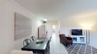 This single-floor residence offers a well-organized layout with light-filled interiors and consistent tile flooring throughout. The open-plan living and dining area forms the heart of the space, where a sectional sofa and a patterned area rug anchor the living zone, complemented by a flat-screen TV on a media console. Crown molding runs along the ceilings, adding a clean architectural detail to the neutral, cream-toned walls. A large window fitted with vertical blinds allows natural light to filter in while maintaining privacy. The dining area features a dark-topped table with mixed seating and is accented by a large abstract wall canvas, giving the space a contemporary touch. Adjacent to the dining area, the kitchen is equipped with open metal shelving, wood-cabinet counters with a dark surface, a refrigerator, and a rolling prep station, offering a practical and organized cooking environment. The primary bedroom includes a metal-frame bed, a generous sliding-door closet with built-in storage, and tile flooring. A secondary room serves as a flexible bedroom or workspace, featuring a wide desk, large windows with vertical blinds providing views of surrounding greenery, and a cozy, functional atmosphere throughout.