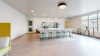This single-floor community room in Madison, WI offers a generously sized, open-plan layout designed to accommodate a variety of social and recreational activities. The space is finished with large-format tile flooring throughout, crisp white walls, and recessed lighting complemented by modern pendant fixtures, creating a bright and welcoming atmosphere.

A standout feature is the long, white marble-topped bar counter, lined with green stools and backed by a bold geometric teal-and-white tile accent wall. A coffee and beverage station adds functional appeal to this central gathering point. Pendant lights hang above the counter, enhancing both form and function.

The lounge area is defined by a wall-mounted flat-screen TV above a natural wood console table, with a round upholstered ottoman providing casual seating. A colorful abstract painting and high-top bistro table with metal barstools add character to this corner. A vibrant yellow-green privacy booth near the entrance offers a semi-enclosed nook for quieter use.

Large windows and glass doors along one wall flood the space with natural light and provide views of the surrounding neighborhood, while double entry doors with glass panels ensure easy accessibility throughout.