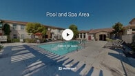 Located at 28155 Encanto Drive in Menifee, California, this outdoor community amenity space centers around a generously sized swimming pool with clear, blue-green water, showcasing a well-maintained and thoughtfully designed recreational environment. The pool is framed by a broad concrete sundeck lined with lounge chairs, ideal for sunbathing under the Southern California sky. Mature shade trees are strategically positioned throughout the area, providing natural relief from the sun while enhancing the overall aesthetic of the space. A decorative pergola with white columns adds architectural interest to one end of the pool area, creating a shaded gathering spot accented by potted plants and additional seating. Tall palm trees contribute to the resort-like atmosphere, reinforcing the warm, Southern California ambiance. Adjacent to the pool, a single-story clubhouse building features stone accents, a pitched tile roof, large windows, and distinctive red double doors, serving as a functional and visually appealing anchor to the space. A separate covered structure with a gazebo-style canopy and red umbrella provides additional outdoor seating options. The entire pool area is enclosed by a wrought iron fence, ensuring privacy and security. Multi-story residential buildings visible in the background complete the community setting.