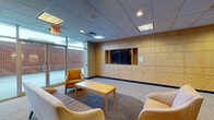 Located at 2100 E High St in Springfield, Ohio, this single-floor facility offers a well-appointed and functional interior designed to accommodate both comfort and practicality. The most prominent area visible is a generously sized lounge or common room, characterized by its warm, neutral tones and a drop ceiling with recessed and fluorescent lighting. A full wall of built-in light wood cabinetry dominates one side of the room, providing extensive storage and housing a centrally mounted flat-screen television — a clear focal point for the space. The lounge is furnished with comfortable upholstered seating, including sofas and an armchair arranged around a wood-topped coffee table, all set upon a patterned area rug over carpeted flooring. Large glass doors along one wall allow natural light to filter in, offering views to the exterior brick building surroundings. Additional rooms within the space appear to be private or utility-oriented, featuring window blinds for light control and minimal furnishings. The overall ambiance of the space is calm and institutional in character, with a clean, organized layout suited for a professional, educational, or community-use setting.