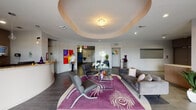 Stadium Tower's ground-floor lobby in Miami's Little Havana neighborhood offers a polished, contemporary welcome experience. The centerpiece of the main lounge is a dramatic circular tray ceiling with a crystal chandelier, creating a defined focal point above a seating area furnished with sleek gray sofas and chrome-framed chairs arranged atop a bold purple peacock-motif area rug. The flooring transitions between warm wood-tone planks and large-format tile, reinforcing distinct functional zones throughout the space. A curved reception desk, accented with pendant lighting and a backlit display, anchors the concierge area and is complemented by a striking blue canvas artwork and a sculptural wall-mounted spider installation, adding a distinctive artistic character. Colorful, large-scale paintings are displayed throughout, lending the lobby a gallery-like atmosphere. Floor-to-ceiling windows allow natural light to penetrate the interior while providing views of the surrounding streetscape. Adjacent to the main lounge, a dedicated mail room features a full bank of resident mailboxes, a central sorting counter, and secured entry doors bearing the Stadium Tower name. The overall design balances modern architecture with vibrant artistic expression, creating a functional yet visually engaging communal space for residents and visitors alike.