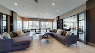 This stylish single-floor community clubhouse is thoughtfully designed to offer residents a variety of inviting social and relaxation spaces. The grand entrance lobby sets a refined tone with large-format marble-look tile flooring, soaring double-height glass walls that flood the space with natural light, exposed brick accent walls, and structural columns that frame the room with architectural elegance. Sleek black-framed glass doors connect the lobby to the rest of the facility, creating a seamless yet defined flow between zones. The main lounge area features generous sectional sofas arranged around low-profile coffee tables, all oriented toward expansive windows overlooking an outdoor pool deck. A coffee bar area showcases a striking exposed brick backdrop, dark granite countertops, built-in shelving with greenery, and a mounted display screen, adding both function and character. A more intimate seating nook offers plush armchairs, floor-to-ceiling draped windows, and dark wood-look tile flooring, creating a quieter retreat. The dining area continues the warm aesthetic with large windows, sheer draping curtains, artful wall panels, and a mix of communal and intimate table settings. Decorative hexagonal tile inlays and curated artwork throughout add personality and visual interest to the overall space.