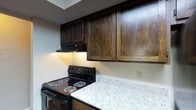 This single-floor space offers a thoughtfully designed interior with a kitchen that serves as a standout feature. The kitchen is outfitted with rich, dark-stained wood cabinetry that extends to the upper walls, providing generous storage while lending a warm, sophisticated character to the space. The cabinetry's deep grain and bold finish create a striking visual contrast against the lighter elements of the room. Below, light-toned granite-style countertops offer ample workspace and contribute a clean, polished aesthetic. A black electric range with coil burners sits seamlessly alongside the countertop, complemented by an under-cabinet range hood that keeps the cooking area practical and well-ventilated. The walls feature a subtle textured finish, adding depth and dimension to the overall design. Recessed under-cabinet lighting provides warm, functional illumination across the countertop workspace, enhancing both usability and ambiance. The kitchen's layout is efficient and purposeful, making the most of the available space. Overall, the interior strikes a balance between classic warmth and modern functionality, with the dark wood cabinetry and stone-look countertops creating a cohesive and inviting environment throughout the single-floor layout.