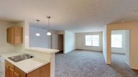 This single-story residence features three bedrooms and two bathrooms within a well-organized floor plan. The open-concept living and dining area is carpeted and bathed in natural light from a large window, creating a bright and welcoming atmosphere. The kitchen includes a raised peninsula countertop with wood-toned cabinetry beneath, providing additional prep space and a casual seating or serving area. Two pendant lights hang above the peninsula, offering focused task lighting while adding a subtle decorative touch. The textured ceilings and neutral wall tones throughout contribute to a clean, understated aesthetic. A closet is visible near the living area, offering convenient storage. The seamless flow between the kitchen and living spaces makes the layout practical for everyday living and entertaining. Overall, the home presents a straightforward, functional design with comfortable finishes suited for a variety of lifestyles.