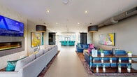 Located at 723 Wheatland St in Phoenixville, PA, this expansive single-floor amenity space offers a dynamic and thoughtfully designed environment for socializing and recreation. The open-plan layout flows seamlessly from zone to zone, anchored by polished concrete floors and bright, neutral walls that serve as a canvas for bold, large-format contemporary artwork throughout.

The main lounge area features generous sectional seating arranged around a sleek linear fireplace with a mounted flat-screen TV above, all illuminated by oversized drum pendant lights. Decorative perforated room dividers add architectural interest while defining separate seating clusters, including a navy blue leather sofa grouping with vibrant accent pillows.

A curved bar anchors the far end of the space, outfitted with teal upholstered barstools and floor-to-ceiling windows that flood the area with natural light and offer views of the outdoor courtyard. A dedicated game room showcases a dramatic full-wall industrial mural, a billiards table, and bar-height seating alongside a living moss wall accent. A secondary activity area includes a foosball table and casual café-style seating with direct glass door access to the exterior courtyard. Throughout, the design balances an energetic, modern aesthetic with comfortable, community-focused functionality.