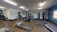 The fitness center at Park At Hurstbourne is a spacious, single-floor facility designed to support a full range of workout routines. The room is finished with durable patterned carpet flooring and illuminated throughout by bright overhead fluorescent lighting, creating a clean and energetic atmosphere. Large windows along one wall invite natural light and provide outdoor views, enhancing the overall environment. The cardio section is well-stocked with Matrix-brand treadmills and elliptical trainers, as well as stationary bikes, each equipped with individual monitor screens. A dedicated strength-training area features a Matrix cable and weight machine, a power tower for bodyweight exercises, and adjustable benches for a variety of resistance workouts. The space also includes a functional open floor zone with medicine balls, resistance accessories, and wall-mounted storage racks for small equipment. A flat-screen television is mounted on the wall for entertainment during workouts. The walls are painted in a combination of light gray and a bold accent blue, adding visual interest to the space. A mirrored wall in the cardio area expands the sense of space. The fitness center is a comprehensive, well-organized amenity suited for both cardiovascular and strength training needs.