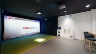 Located at 1140 E Market St in Akron, Ohio, this indoor golf simulator bay offers a fully equipped, self-contained experience for golf enthusiasts of all skill levels. The single-room space is thoughtfully designed with two distinct zones that work seamlessly together. The centerpiece is a TruGolf simulator setup, featuring a large projection screen framed by dark-paneled walls and accent lighting, creating an immersive atmosphere. A synthetic turf hitting mat is positioned directly in front of the screen, providing a realistic feel underfoot during play. A ceiling-mounted projector delivers high-definition simulation software, supporting both practice modes and full course play via the e6 Golf platform. Adjacent to the simulator area, a comfortable spectator and waiting zone features a wall-mounted wooden bar counter lined with modern bar-height chairs, perfect for guests to relax and watch. Black-and-white golf photography adorns the white walls, adding a classic sporting character to the space. A dedicated equipment station houses a monitor, club storage, and a collection of practice balls and various sports equipment. Recessed lighting illuminates the seating area, while the dark ceiling overhead reinforces the dramatic, focused ambiance of the simulator bay. The space connects directly to an adjacent lounge area, enhancing the overall social experience.