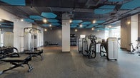 Located at 600 North Beach on Atlantic Avenue in Atlantic City, New Jersey, this fitness center offers a well-appointed, single-floor exercise facility designed with both functionality and aesthetics in mind. The space features an industrial-style exposed ceiling finished in dark tones, accented by striking teal hexagonal acoustic panels that add visual interest while managing sound. Track lighting and pendant fixtures illuminate the floor evenly throughout.

The main workout area is divided into distinct functional zones. The strength training section houses a comprehensive selection of Technogym resistance machines, a cable cross machine, adjustable benches, and a rack of dumbbells. A dedicated free-weight and functional training area features a smooth hardwood floor, ideal for stretching, rowing, and medicine ball work. A well-stocked rack of medicine balls and stability balls lines the textured gray accent wall.

The cardio zone, positioned near large windows that invite natural light, is equipped with a row of treadmills, stationary bikes, elliptical trainers, and stair climbers, offering views to the exterior. Durable carpet tiles cover the primary workout floor, complementing the hardwood sections. The facility is accessed via modern interior doors and is clearly marked with emergency exits, ensuring a safe and well-organized environment for users.