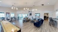 This single-floor communal space is designed to offer both comfort and functionality across several distinct activity zones. The interior is characterized by soaring high ceilings that amplify the sense of openness, complemented by large glass doors that allow generous amounts of natural light to flood the space and provide views of the exterior surroundings. The flooring throughout is a warm-toned hardwood-style plank, lending a cohesive and contemporary feel to the entire area. Multiple statement chandeliers with a modern sputnik-inspired design hang overhead, adding a stylish focal point to the lounge area. Comfortable seating is arranged thoughtfully, featuring a gray sofa paired with a round wooden coffee table and a set of rich navy blue accent chairs, creating an inviting social hub. A wall-mounted flat-screen television is positioned nearby, enhancing the entertainment options. In the foreground, a shuffleboard table adds a recreational element, while toward the rear, large glass doors open to a visible fitness area equipped with exercise equipment. Bar-height tables and stools near the windows offer additional casual seating. The overall ambiance is bright, modern, and welcoming.