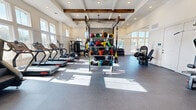 This impressive fitness center combines elegant architectural design with a full suite of professional-grade exercise equipment. The space is defined by its soaring ceilings adorned with rustic wood beams set within coffered detailing, creating a striking visual contrast against the bright white walls and recessed lighting. An abundance of natural light pours in through expansive floor-to-ceiling windows lining multiple walls, offering outdoor views and an open, airy atmosphere throughout.

The facility is organized into distinct training zones. A dedicated cardio area features multiple treadmills and stationary cycling bikes, including ICG-branded spin bikes, all positioned to take advantage of the natural light. A functional training section showcases a large RAX multi-purpose rig equipped with TRX suspension trainers, a punching bag, battle ropes, and an extensive rack of colorful medicine balls and resistance accessories. The strength training area includes a full dumbbell rack, adjustable benches, cable machines, and a multi-station weight apparatus, complemented by floor-to-ceiling mirrors that enhance the sense of space. Durable rubber flooring spans the entire facility, providing safety and comfort underfoot. An open stretch and flex area with additional mirrors rounds out this comprehensive, well-appointed fitness space.
