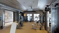 This fitness center offers a comprehensive workout environment designed with both functionality and aesthetics in mind. The space features a vaulted ceiling with warm wood-paneled accents and recessed lighting, complemented by a central ceiling fan that promotes air circulation throughout. The walls incorporate a striking stacked stone accent detail framing the entryway, adding a refined, modern-rustic character to the room. Large windows along the far wall flood the interior with natural light and provide an open, airy feel. The flooring is finished in a neutral carpet that softens the space while providing comfort underfoot. Equipment is thoughtfully arranged throughout, including a variety of strength-training machines and multi-use cable systems positioned along the perimeter, alongside a selection of cardio equipment such as ellipticals, stationary bikes, and a treadmill near the windows. Wall-mounted mirrors enhance the sense of space and allow users to monitor their form. Decorative sconce lighting adds warmth alongside the functional overhead fixtures. The overall ambiance strikes a balance between a polished, upscale aesthetic and a practical, fully equipped athletic facility suitable for a range of fitness routines.