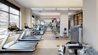 This modern fitness center is a comprehensively outfitted facility designed to support a wide range of workout routines. The space is organized into distinct zones, with a dedicated cardio area lining the windowed perimeter and a robust strength training section occupying the central and rear portions of the floor.

The cardio zone features a long row of treadmills, stair climbers, elliptical machines, and rowing ergometers, all positioned along large, floor-to-ceiling windows that invite abundant natural light and provide open views of the surrounding area. Mounted flat-screen televisions are positioned throughout for entertainment during workouts.

The strength training area is stocked with a wide selection of cable machines, selectorized weight stations, free weight benches, and a full dumbbell rack, all arranged to allow for efficient circulation. Large mirrors line the walls, enhancing the sense of openness and allowing users to monitor their form.

The flooring throughout is a durable, cushioned material well-suited to high-traffic fitness use. Overhead, linear LED lighting ensures the entire space is evenly and brightly illuminated. A decorative navy blue accent wall with an abstract wave motif adds a contemporary design touch. A glass-enclosed room near the entrance, with posted hours, provides an additional private workout or studio space. The overall ambiance is clean, professional, and well-maintained.