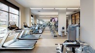 This modern fitness center is a comprehensively outfitted facility designed to support a wide range of workout routines. The space is organized into distinct zones, with a dedicated cardio area lining the windowed perimeter and a robust strength training section occupying the central and rear portions of the floor.

The cardio zone features a long row of treadmills, stair climbers, elliptical machines, and rowing ergometers, all positioned along large, floor-to-ceiling windows that invite abundant natural light and provide open views of the surrounding area. Mounted flat-screen televisions are positioned throughout for entertainment during workouts.

The strength training area is stocked with a wide selection of cable machines, selectorized weight stations, free weight benches, and a full dumbbell rack, all arranged to allow for efficient circulation. Large mirrors line the walls, enhancing the sense of openness and allowing users to monitor their form.

The flooring throughout is a durable, cushioned material well-suited to high-traffic fitness use. Overhead, linear LED lighting ensures the entire space is evenly and brightly illuminated. A decorative navy blue accent wall with an abstract wave motif adds a contemporary design touch. A glass-enclosed room near the entrance, with posted hours, provides an additional private workout or studio space. The overall ambiance is clean, professional, and well-maintained.