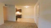 This single-floor residential unit offers a straightforward and functional layout designed for comfortable everyday living. The open-concept design allows the living area and kitchen to flow naturally into one another, creating a cohesive and practical space.

The living area features wall-to-wall carpeting in a neutral beige tone, providing a warm and understated foundation for the room. Smooth, light-colored walls and a flush ceiling light fixture contribute to a clean, simple ambiance throughout the space. A door on the far wall leads to an adjacent room, offering additional privacy and separation within the unit.

The kitchen is positioned at the rear of the open living space and is equipped with dark wood cabinetry that provides a subtle contrast against the lighter walls and flooring. The cabinetry offers both upper and lower storage, accompanied by a countertop workspace and a sink with under-cabinet lighting. The kitchen floor transitions to a lighter-toned hard surface, clearly delineating the cooking area from the carpeted living zone.

Overall, the space presents a no-frills, move-in-ready environment with a calm, neutral palette. The open layout maximizes the sense of space, while the practical finishes make it easy to personalize and maintain.