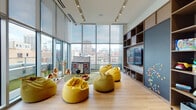 This thoughtfully designed children's amenity space combines playful creativity with modern architecture to deliver an engaging, welcoming environment. Soaring floor-to-ceiling glass curtain walls flood the room with abundant natural light while framing expansive panoramic city views, creating a vibrant backdrop that sets the space apart. Light-toned hardwood flooring runs throughout, lending warmth and continuity to the interior.

The main play area is anchored by a circular green area rug, where a charming miniature log cabin playhouse serves as a focal point alongside whimsical toy animals and decorative tree accents. Plush, mustard-yellow bean bag chairs are scattered throughout, offering comfortable and casual seating for children of all ages. A full-height built-in shelving and media unit lines one wall, featuring integrated storage cubbies, a flat-screen television, and a magnetic chalkboard panel — ideal for interactive and creative play.

Adjacent to the main lounge is a transitional lounge nook finished with warm wood-toned wall paneling, stylish globe wall sconces, terrazzo-topped café tables, and upholstered bench seating — providing a comfortable retreat for supervising adults. A concrete utility sink nearby adds practical functionality to the space. Together, these areas form a cohesive, well-appointed amenity designed for both play and relaxation.