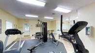 This single-floor fitness space offers a practical and well-organized environment for exercise. The room is illuminated by multiple large flush-mounted fluorescent light panels in the ceiling, ensuring consistent, bright lighting throughout the space. The walls are painted in a soft yellow tone, lending a warm and welcoming atmosphere to the room.

The flooring features a checkered tile pattern in neutral tones, adding visual interest while remaining durable and easy to maintain. A multi-station cable machine serves as the centerpiece of the room, accompanied by a weight bench, a treadmill, and additional cardio equipment, providing a well-rounded range of workout options.

Along the back wall, a row of what appears to be laundry machines or storage units is visible, suggesting a dual-purpose utility area adjacent to the fitness space. A windowed door on the left side of the room allows natural light to filter in, offering a view of the outdoor surroundings. Framed artwork decorates the walls, adding a touch of character. Overall, the space is clean, functional, and thoughtfully arranged to serve the fitness needs of its users.
