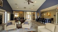 This single-floor space offers a thoughtfully designed open-concept layout that seamlessly integrates multiple functional areas into one cohesive environment. The lounge area anchors the space with comfortable cream-upholstered armchairs arranged around a round side table, set atop a patterned area rug that adds texture and warmth. Decorative columns frame the open floor plan, lending a classic architectural character that is complemented by crown molding and bold blue-gray accent walls.

Natural light filters in through glass-paneled entry doors, brightening the central corridor and contributing to the airy atmosphere. Recessed lighting and a ceiling fan with integrated lighting ensure the space remains well-lit and comfortable throughout. Colorful artwork adorns the walls, adding visual interest and a curated aesthetic.

Toward one side, a dedicated workstation area features dark wood desks and task chairs, making it suitable for business or administrative use. On the opposite side, a bar-height counter with dark dining chairs serves as a casual dining or refreshment station, accompanied by pendant lighting that adds a modern touch. The combination of tile and area rug flooring throughout delineates each zone while maintaining a unified, polished look across the entire space.