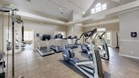 Located at 2101 21st St SE in Hickory, NC, this single-floor fitness center offers a comprehensive workout facility spread across a generously sized footprint. The space is organized into three distinct areas, each designed to support a range of fitness activities.

The main workout hall is the architectural centerpiece, boasting soaring vaulted ceilings with a clerestory arched window that floods the space with natural light. Track lighting runs along the ceiling, complementing the bright, open atmosphere. Large French doors provide outdoor access and further enhance the sense of openness. This primary area houses an array of cardio machines, including treadmills and elliptical trainers, as well as a cable weight machine, all set atop durable tile flooring with crown molding accents throughout.

An adjoining strength training room features rubberized flooring, a full-length mirror, ample natural light from multiple windows with blinds, and a well-organized selection of free weights, barbells, and a cable/weight machine — suited for a thorough strength workout.

A third room serves as a flexible exercise space with rubberized flooring, a wall-mounted television, and access to the main hall via French doors, making it ideal for stretching, mat work, or cardio equipment use. The consistent warm neutral tones, crown molding, and abundant natural light throughout create a clean, welcoming environment for fitness activities.