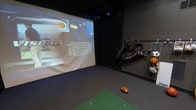 This single-floor sports simulation room is purpose-built for immersive athletic entertainment, combining cutting-edge technology with thoughtful design. The room is finished in deep, dark tones — walls and ceiling are painted black to eliminate glare and enhance the visual experience of the large-format projection screen that dominates one wall. The screen serves as the centerpiece of the space, displaying a multi-sport simulation interface capable of supporting golf, basketball, and other virtual sporting experiences, with precision shot-tracking technology built into the system.

The flooring transitions from a synthetic green turf hitting mat — complete with tee markers — to a hard floor area, providing an authentic feel for golf swings and other sport activities. A football rests casually on the turf, nodding to the room's multi-sport versatility. Along the side wall, a well-organized golf bag rack holds a full set of clubs and a golf bag, keeping equipment accessible and neatly stored.

Recessed and track lighting overhead provides focused illumination without interfering with the projected display. The overall ambiance is that of a professional-grade simulator bay — intimate, focused, and fully equipped for serious practice sessions or recreational play.