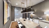 This professionally designed conference room offers a refined and functional environment ideal for meetings and collaborative sessions. At the heart of the space sits a long, light-toned rectangular meeting table surrounded by sleek white upholstered chairs with caster bases, comfortably accommodating a sizable group of attendees. Overhead, a pair of dramatic rectangular pendant light fixtures finished in matte black and brushed gold hang from the ceiling, providing focused illumination while serving as a bold design statement. The walls are finished in a deep charcoal tone, lending the room a contemporary and professional ambiance. A standout architectural feature is the full-height glass partition wall adorned with an intricate geometric patterned overlay, which visually separates the room from an adjacent space while allowing light to filter through. Warm terracotta-toned flooring adds an earthy contrast to the otherwise cool, modern palette. A built-in credenza along one wall provides additional surface space and storage, complemented by a table lamp with a gold base that adds a warm accent. Recessed ceiling lighting supplements the pendant fixtures, ensuring the room is well-lit throughout. The overall design balances elegance with functionality, creating a polished setting for professional use.