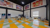 This energetic recreation space is designed to entertain and engage visitors of all ages. The interior immediately captures attention with its dramatically high ceilings framed by bold black structural beams and a dark ceiling, creating a striking industrial-modern aesthetic. Lining the upper walls is an extensive collection of large-format pop-art and video game-inspired murals, featuring iconic imagery that adds a playful, gallery-like atmosphere throughout the room.

The floor is a standout design element, composed of a vibrant checkerboard pattern in yellow, green, white, and orange tiles that energizes the entire space. Natural light filters in through multiple windows fitted with layered roller blinds, while large glass doors at one end provide views of an outdoor playground area beyond.

The space is thoughtfully divided into activity zones. A gaming area houses classic arcade cabinets, a foosball table, and an air hockey table, alongside racing-style gaming chairs and wall-mounted flat-screen TVs. A relaxed seating zone offers sofas and lounge chairs for social gathering, while a separate dining area provides multiple tables and chairs for casual meals or group activities. Vending machines are also conveniently accessible. The open, airy layout and cohesive playful design make this a dynamic and inviting recreational destination.