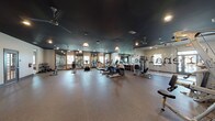This single-floor fitness center offers a spacious, open layout designed to accommodate a comprehensive workout experience. The interior is defined by a striking dark ceiling that contrasts with light-toned walls, creating a focused and energizing atmosphere. Recessed lighting is complemented by decorative pendant fixtures, providing well-distributed illumination throughout the space.

The main exercise area is generously proportioned, featuring durable rubberized flooring that supports high-impact activity while providing comfort underfoot. Large mirrors line multiple walls, enhancing the sense of openness and allowing users to monitor their form during workouts.

The cardio section is well-appointed with multiple treadmills, elliptical machines, and stair climbers arranged to allow ample space between each unit. The strength training area includes a multi-function cable machine suitable for a variety of resistance exercises, as well as a rack of free weights for versatile training options. Storage cubbies provide convenient organization for equipment and personal items.

Natural light filters in through several windows fitted with blinds, adding to the bright and welcoming environment. Ceiling fans support air circulation, keeping the space comfortable during intense workouts. Glass-panel doors with dark frames provide a polished entryway, and safety features such as clearly marked emergency exits are present throughout. Overall, the fitness center is thoughtfully designed for both function and aesthetic appeal.