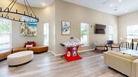 Located at 1 Royal Dr in Somersworth, New Hampshire, this single-floor community space offers a bright and welcoming environment designed for both relaxation and social engagement. The open-concept layout is anchored by a modern kitchen outfitted with stainless steel appliances — including a full-size refrigerator with ice maker, electric range, and built-in microwave — white cabinetry, a clean white countertop, and a two-tone island with bar seating in bold red chairs. Light wood-style flooring runs throughout, complementing the neutral beige and white walls that lend the space a fresh, airy feel. The living area transitions seamlessly from the kitchen, offering comfortable seating arrangements beneath recessed lighting and a decorative chandelier. A large wall-mounted flat-screen TV serves as a focal point for gatherings, while mid-century modern accent chairs add style and function. The game area is a standout feature, complete with a tabletop bubble hockey game, a mounted Scrabble board with chalkboard, and a bistro-style high-top table. New Hampshire-themed artwork adds local character throughout. Two hallways connect the zones efficiently, and multiple windows invite abundant natural light while framing views of the surrounding greenery.