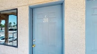 Unit C201 is identified by its distinctive light blue six-panel entry door, accented with silver unit numbers against a textured stucco exterior facade. The single-floor layout is designed with an open-concept approach, seamlessly integrating the living, sleeping, and kitchen areas into one cohesive space.

The main living area features soft, neutral-toned carpet underfoot and light blue walls that create a calm, airy atmosphere throughout. A comfortable sleeping area occupies the foreground of the space, benefiting from the open sightlines. The room transitions naturally into a defined dining nook, which accommodates a small table and chairs beneath a pendant light fixture, providing an intimate spot for meals.

The kitchen is partially separated by a subtle architectural division, offering a sense of distinction without fully closing off the space. Warm wood-toned cabinetry lines the kitchen walls, complemented by standard appliances including a range and oven. Recessed and overhead lighting illuminate the kitchen area effectively.

Overall, the unit maintains a cohesive, light palette throughout, with consistent flooring and wall tones that enhance the sense of openness across the entire space.