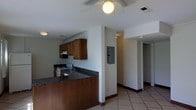 This single-floor space is designed with a practical, open layout that connects the kitchen and living areas seamlessly. The kitchen is a standout feature, showcasing dark granite countertops that contrast with warm wood cabinetry, providing ample storage space. A white electric range and under-cabinet ventilation hood complete the cooking area, while a sink is conveniently positioned along the countertop. The kitchen extends into a peninsula-style counter, which serves as a natural divider between the cooking and living spaces. Light beige ceramic tile flooring runs throughout, offering durability and easy maintenance. A ceiling fan with a light fixture is centrally positioned, ensuring both air circulation and overhead lighting. Visible HVAC vents are integrated into the ceiling for climate control. An electrical panel is neatly recessed into the wall in the transitional hallway area, keeping utilities accessible yet unobtrusive. The hallway leads toward additional rooms, with clean white walls and consistent tile flooring maintaining a cohesive aesthetic throughout. The overall ambiance is functional and straightforward, well-suited for everyday living.