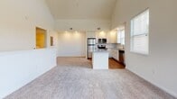 Located at 100 Shellbark Way in Savannah, GA 31407, this well-appointed home offers a thoughtfully designed single-floor layout with standout architectural details. The open-concept living and kitchen area is anchored by soaring vaulted ceilings that create an impressive sense of volume and airiness, with natural light streaming in through multiple windows fitted with blinds. The kitchen is a functional centerpiece, featuring white cabinetry, granite countertops, a central island, and a full suite of stainless steel appliances including a refrigerator, range, microwave, and dishwasher. Rich hardwood flooring runs through the kitchen, while plush carpet covers the living area and bedrooms. The primary bathroom offers a spacious layout with white cabinetry, dark countertops, a large mirror, and hardwood flooring, complemented by a walk-in closet and separate water closet. Additional bedrooms are carpeted and filled with natural light through bright windows. A dedicated laundry area houses a full-size washer and dryer with overhead shelving for added storage. The staircase, lined with a warm wood handrail and carpeted treads, provides access to the upper loft-style space visible from the main level. Overall, the home presents a clean, move-in-ready ambiance with modern conveniences throughout.