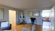 This single-floor space offers a well-organized layout that balances open communal areas with private living quarters. The living room serves as the central gathering space, featuring warm hardwood flooring, neutral gray walls, and clean white trim. Framed artwork adorns the walls, complementing a sleek media console and a large floor mirror that adds depth and brightness to the room. A comfortable seating arrangement anchors the space, which flows naturally into the adjacent areas.

The kitchen is positioned toward the rear of the unit, offering functional counter space and cabinetry. Natural light filters through windows, creating a bright and airy atmosphere throughout the shared living and kitchen zone.

A separate wing accommodates the bedroom and bathroom, providing a degree of privacy from the main living areas. The bathroom features tiled flooring, distinguishing it from the hardwood found throughout the rest of the space.

Overall, the layout is compact yet efficient, with consistent finishes — including the warm hardwood flooring and neutral color palette — tying the entire space together into a cohesive and inviting environment.