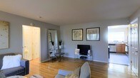 This single-floor space offers a well-organized layout that balances open communal areas with private living quarters. The living room serves as the central gathering space, featuring warm hardwood flooring, neutral gray walls, and clean white trim. Framed artwork adorns the walls, complementing a sleek media console and a large floor mirror that adds depth and brightness to the room. A comfortable seating arrangement anchors the space, which flows naturally into the adjacent areas.

The kitchen is positioned toward the rear of the unit, offering functional counter space and cabinetry. Natural light filters through windows, creating a bright and airy atmosphere throughout the shared living and kitchen zone.

A separate wing accommodates the bedroom and bathroom, providing a degree of privacy from the main living areas. The bathroom features tiled flooring, distinguishing it from the hardwood found throughout the rest of the space.

Overall, the layout is compact yet efficient, with consistent finishes — including the warm hardwood flooring and neutral color palette — tying the entire space together into a cohesive and inviting environment.
