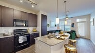 This single-floor residence offers a thoughtfully designed open-concept layout that seamlessly connects the kitchen, dining, and living areas. The kitchen is a standout feature, showcasing rich dark espresso cabinetry, granite countertops with a subway tile backsplash, and a full suite of black appliances including a side-by-side refrigerator, electric range, and built-in microwave. A central island with a double stainless steel sink and pendant lighting overhead serves as both a functional workspace and a casual dining spot, complemented by bar-height seating. Track lighting illuminates the kitchen, while wood-look flooring extends throughout the main living areas, adding warmth and continuity. The living room is bright and welcoming, benefiting from large windows that invite natural light and a ceiling fan that enhances comfort. A private entry door with a transom window further contributes to the airy feel of the space. The bedroom is a calm retreat, carpeted for comfort, featuring a ceiling fan and a generously sized window with blinds. The bathroom is well-equipped with a granite-topped vanity, dark cabinetry, wood-look flooring, and a full soaking tub with a shower combination. A tall linen storage cabinet adds practical storage to the space.