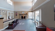 This single-story community space combines functional amenities with an inviting atmosphere across an open, well-appointed layout. The main gathering area commands attention with dramatically vaulted ceilings and clerestory windows positioned high on the walls, flooding the interior with abundant natural light. A striking stone fireplace serves as a central focal point, framed by warm taupe walls, white crown molding, and smooth light-toned flooring that runs throughout.

Large sliding glass doors and expansive windows offer direct views of an outdoor pool area, seamlessly connecting the interior to the exterior. The reception area features a curved, built-in desk finished in complementary warm tones, accompanied by framed certificates and artwork on the walls, conveying a professional yet welcoming environment. A separate seating lounge includes rich dark leather furniture, wall sconces, a decorative mirror, and a console table, creating a comfortable, intimate setting. A glass-top dining table with ornate metal chairs adds a stylish casual seating option nearby. A water cooler station provides added convenience. Access to a fitness center is visible through interior doorways, rounding out the practical offerings of this well-designed communal space.